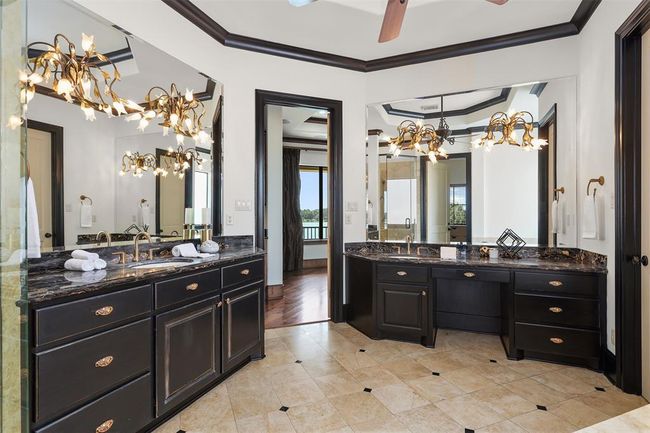 This elegant Primary bathroom features dual vanities with dark cabinetry and luxurious black marble countertops. The space is accentuated by ornate light fixtures, large mirrors, and a neutral tiled floor. | Image 36