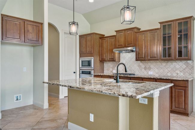 Kitchen featuring light stone counters, arched walkways, backsplash, vaulted ceiling, and brown cabinets | Image 19