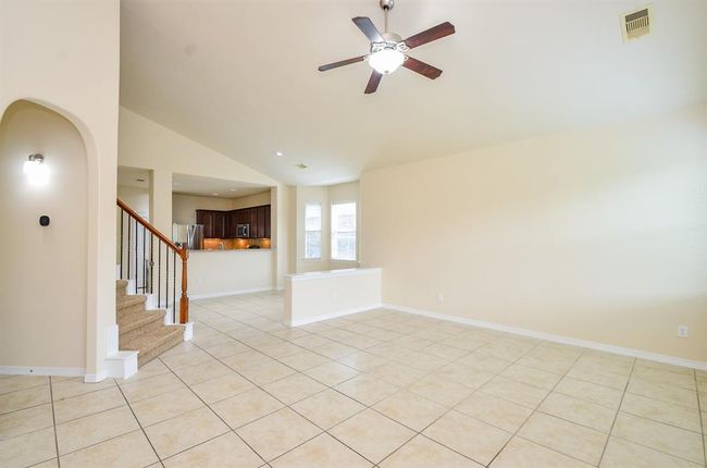 Spacious living area with tiled flooring, high ceiling, and ceiling fan. Features an open layout leading to a modern kitchen with stainless steel appliances. Carpeted stairs add warmth and lead to the upper level. Abundant natural light from large windows. | Image 15