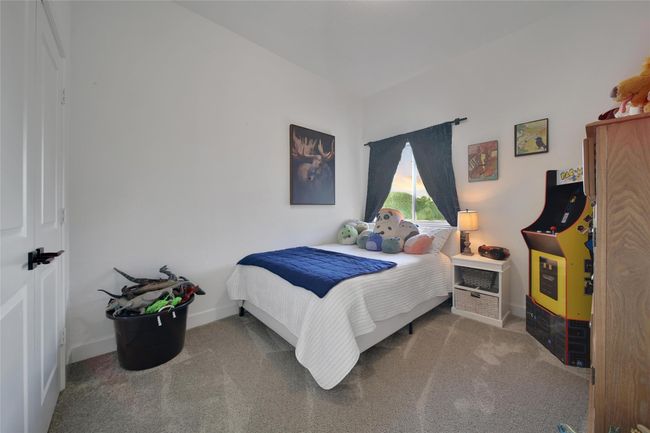 This is a cozy bedroom with a single bed, plush toys, and a small arcade game. The room features a window with dark curtains, a lamp on a nightstand, and carpeted flooring, creating a comfortable and inviting space. | Image 37