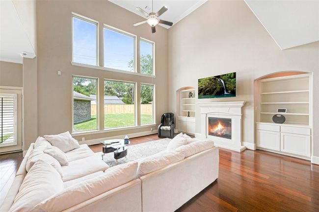 Light-filled living room with soaring two-story ceilings, full wall of windows, and a cozy fireplace flanked by built-in shelving. Offers a seamless view of the backyard and a welcoming space for relaxing or entertaining. | Image 20