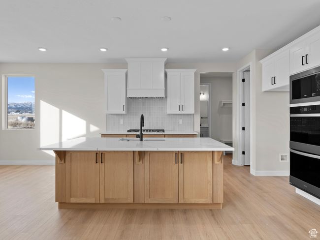 Kitchen with a center island with sink, custom range hood, light countertops, light wood-type flooring, and white cabinetry | Image 10