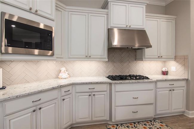 This kitchen features elegant white cabinetry with brushed nickel hardware, a modern stainless steel microwave and hood, a gas cooktop, and a stylish herringbone tile backsplash. The granite countertops offer ample workspace, complemented by under-cabinet lighting for a bright and inviting atmosphere. | Image 16