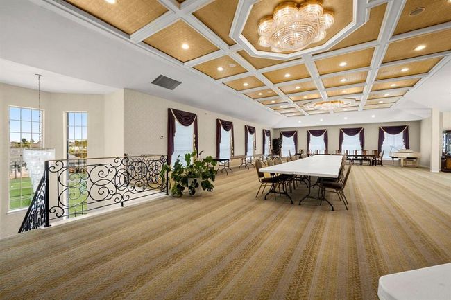 This spacious, well-lit banquet hall features an elegant coffered ceiling with modern chandeliers, large windows dressed with deep purple draperies, and a neutral carpeted floor. It offers a grand setting ideal for hosting events or gatherings, complete with a decorative wrought-iron railing and a view of the outdoor area. | Image 23