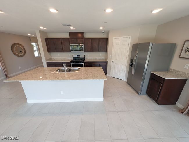 Kitchen featuring stainless steel appliances, dark brown cabinetry, an island with sink, light stone counters, and recessed lighting | Image 8
