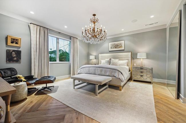 Some primary bedrooms are too oversized, others too small, others, like this one, are JUST RIGHT. A relaxing soft grey blue paint color compliments the decor. This elegant primary features rich herringbone wood flooring, a stunning crystal chandelier, and designer finishes throughout. A serene retreat with ample natural light, cozy sitting area, and a luxurious ambiance perfect for rest and relaxation. | Image 16
