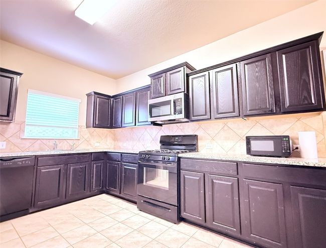 Kitchen featuring black appliances, tasteful backsplash, light tile patterned flooring, light stone counters, and dark brown cabinetry | Image 6