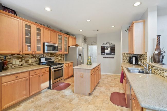 This kitchen features warm wood cabinetry, a central island, stainless steel appliances, and a stylish tile backsplash. The room is well-lit with recessed lighting and offers ample counter space and storage, perfect for cooking and entertaining. | Image 13