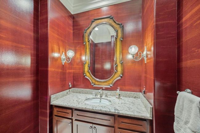 This bathroom features rich wood-paneled walls, a granite countertop, and an ornate oval mirror. It includes elegant wall sconces and a stylish vanity with ample storage. | Image 15