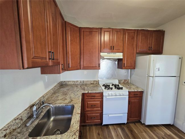 Kitchen with white appliances, under cabinet range hood, dark wood-style flooring, light stone counters, and a sink | Image 6