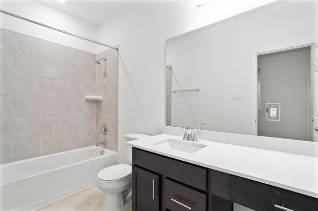 Secondary bathroom features stained cabinets, neutral paint, shower/tub combo with tile surround, large mirror, tile floors, and sleek fixtures and modern finishes. | Image 6