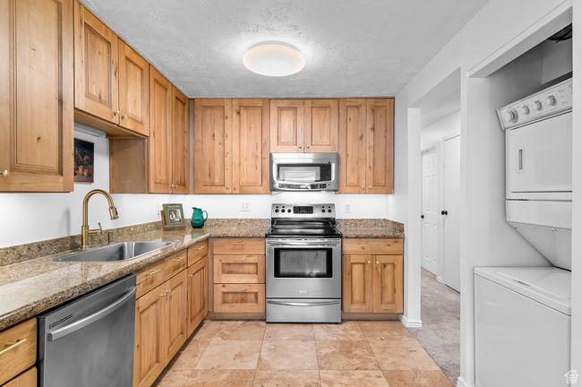 Kitchen featuring stainless steel appliances, stacked washer and clothes dryer, stone countertops, a textured ceiling, and brown cabinetry | Image 5