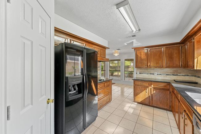 Spacious kitchen with ample natural light, featuring wooden cabinetry, a black refrigerator, and a tiled floor. Adjacent dining area has large windows overlooking the backyard. | Image 12