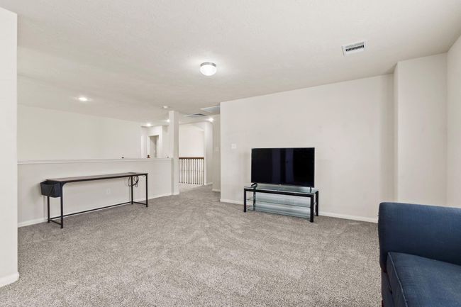 This room is a spacious, carpeted living area with neutral walls, featuring a TV setup and a small table. It has ample lighting and an open layout leading to a hallway, ideal for relaxation and entertainment. | Image 20