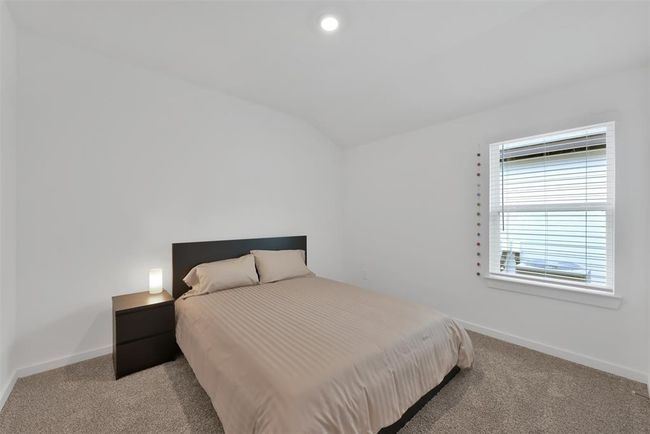 This photo shows a cozy, minimalist bedroom with neutral tones. It features a comfortable bed with a simple headboard, a bedside table with a lamp, soft carpeting, and a window with blinds, allowing natural light to fill the room. | Image 15