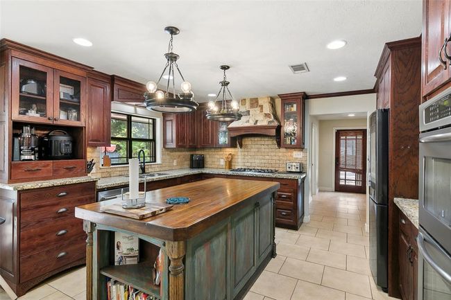This kitchen features rich wooden cabinetry, granite countertops, and a large central island with a rustic finish. It includes modern appliances, a gas stove with a decorative stone backsplash, and stylish pendant lighting, creating a warm and inviting space. | Image 19