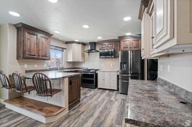 Kitchen with a peninsula, appliances with stainless steel finishes, a kitchen bar, light wood-type flooring, and wall chimney exhaust hood | Image 7