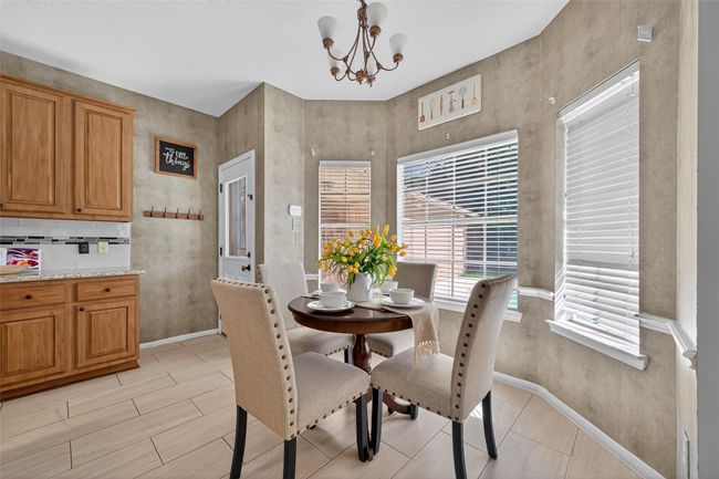 The photo showcases a cozy dining nook with a round table and four upholstered chairs, set against a backdrop of warm, textured walls. Natural light streams in through large windows with white blinds, while wooden cabinetry and a modern light fixture add character. Perfect for intimate meals or morning coffee. | Image 17