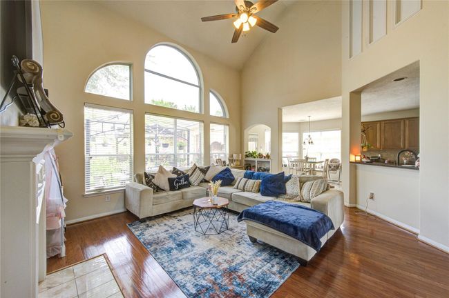 This inviting living room features high ceilings with a ceiling fan, large arched windows for ample natural light, and an open layout that flows seamlessly into the dining area. The cozy seating area includes a modern sectional and a stylish area rug on polished hardwood floors, offering a perfect space for relaxation and entertaining. | Image 17