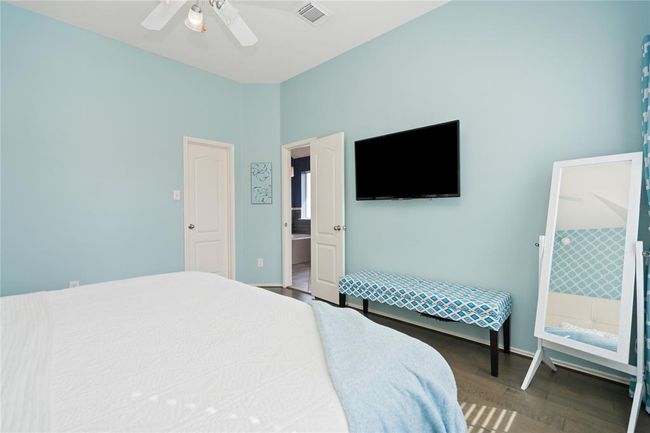 Bright bedroom with light blue walls, featuring a ceiling fan, wall-mounted TV, and a patterned bench. It includes a full-length mirror and an en-suite bathroom. | Image 17