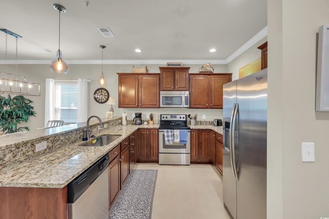 Kitchen with appliances with stainless steel finishes, a peninsula, crown molding, light stone countertops, and brown cabinets | Image 7