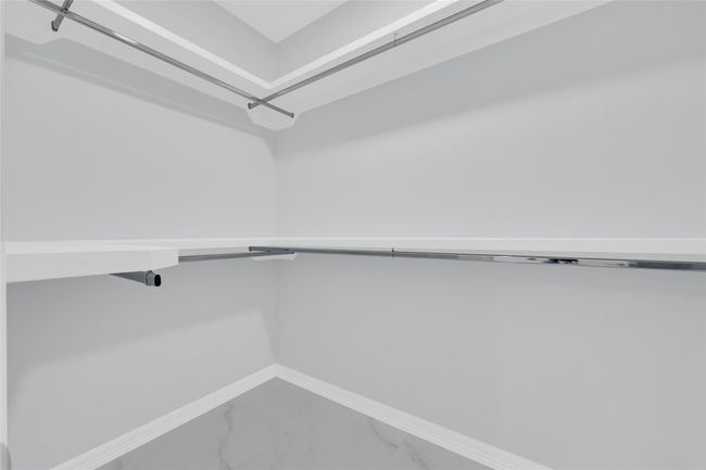 This photo shows a spacious walk-in closet with ample hanging space, featuring sleek chrome rods and a minimalist design. The closet has a clean, modern look with light walls and flooring. | Image 31