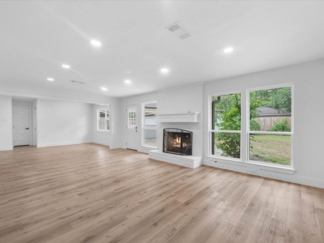 This spacious living room features light wood flooring, a cozy white brick fireplace, and large windows offering plenty of natural light and views of the backyard. The open layout provides ample space for various seating arrangements. | Image 7