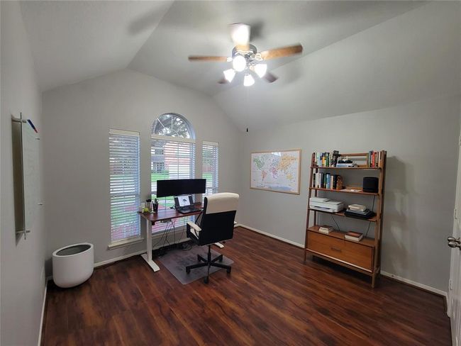 Well-lit home office with a vaulted ceiling and wood flooring. It features a large window and ceiling fan. | Image 26