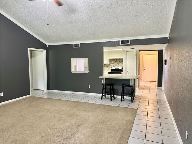 This inviting living space features a combination of carpet and tile flooring, with dark accent walls creating a modern contrast. A breakfast bar with seating connects to the kitchen, which includes stainless steel appliances and a stylish backsplash. The room is spacious with high ceilings, offering ample natural light. | Image 6