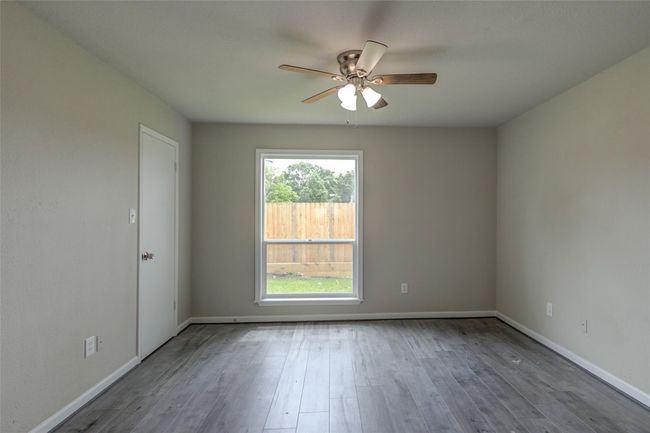 This bedroom features neutral-toned walls and wood-look flooring, creating a modern and inviting space. It includes a ceiling fan for comfort and a large window offering plenty of natural light and a view of the fenced backyard. | Image 13