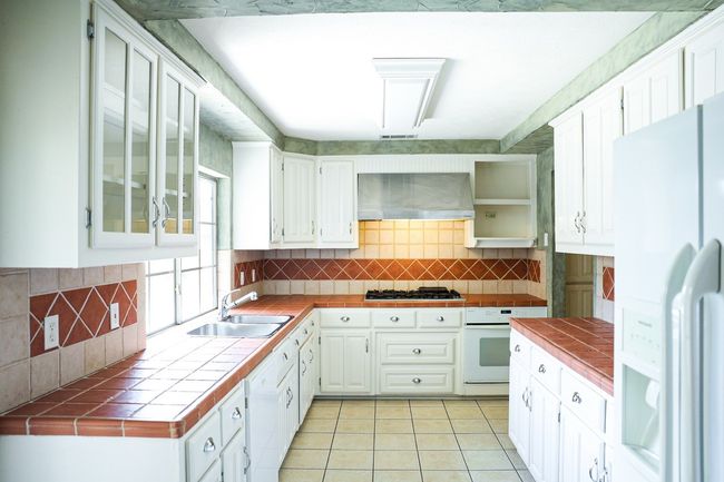 Kitchen with white appliances, tile countertops, wall chimney range hood, and white cabinetry | Image 14