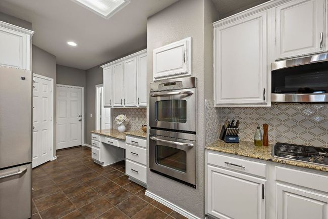 Kitchen featuring appliances with stainless steel finishes, double oven, decorative backsplash, white cabinetry, light stone countertops, and dark tile patterned flooring | Image 15