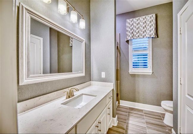 This bathroom features a modern design with a large framed mirror, elegant light fixtures, and a sleek marble countertop. It includes a spacious vanity, stylish tile flooring, and a window with a decorative valance, enhancing natural light. | Image 13