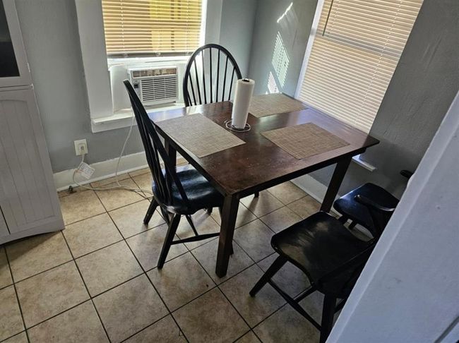 This photo shows a cozy dining area with a dark wooden table and four chairs. The room has tiled flooring, light gray walls, and two windows with blinds, one of which has a window-mounted air conditioning unit. | Image 16