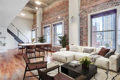 Step into Unit 9B, where natural light floods in through expansive windows, illuminating the open living space. The brick wall adds character and charm, creating a cozy atmosphere for you and your guests. With its high ceilings, this living room feels spacious and inviting, perfect for relaxing or entertaining. This photo is virtually staged. | Image 2