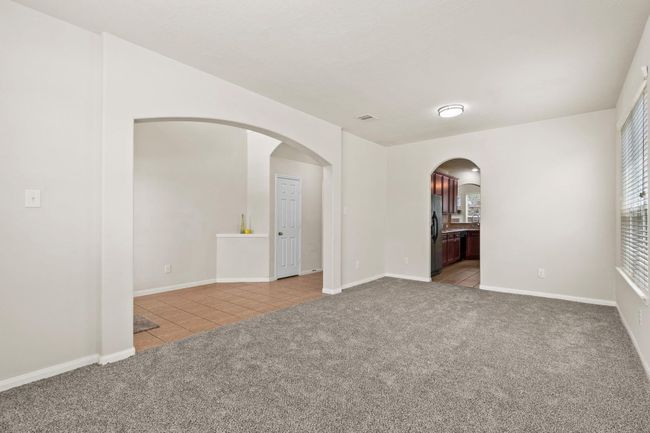 This photo showcases a bright, open living space with neutral walls and carpeting. It features arched doorways leading to a tiled area and a modern kitchen with dark cabinetry. Large windows provide ample natural light. | Image 7