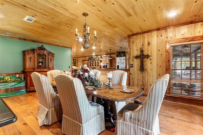 This photo showcases a cozy dining area with wood-paneled walls and ceiling, featuring a rustic chandelier and an elegant dining table with upholstered chairs. The room has a warm, inviting atmosphere, complemented by a vintage-style cabinet and window with wooden blinds. | Image 14