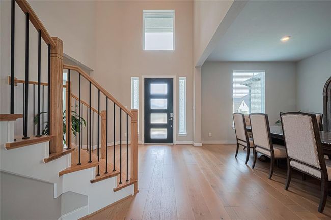 A bright, open entryway with high ceilings w/ modern design. A stylish staircase with wood and iron railing, a contemporary front door with glass panels, and an adjacent dining area with large windows, providing ample natural light. | Image 5
