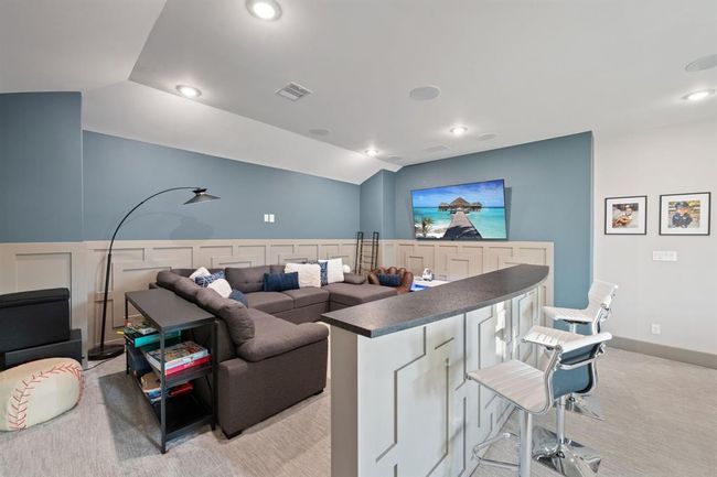 This cozy entertainment room features a large sectional sofa facing a wall-mounted TV, perfect for movie nights. The space includes a modern bar area with seating, ample lighting, and a clean, stylish design with blue and beige tones. Ideal for relaxation and social gatherings. | Image 33