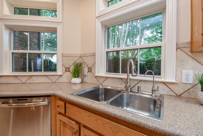 This kitchen features a dual sink with a modern faucet, granite countertops, and a tile backsplash. It has ample natural light from large windows and comes with stainless steel appliances, including a dishwasher. | Image 15