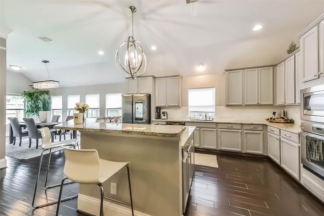 This spacious and modern kitchen features sleek cabinetry, stainless steel appliances, and a large island with seating. The open layout flows seamlessly into a bright dining area, enhanced by natural light from multiple windows. Elegant lighting fixtures add a touch of sophistication. | Image 15