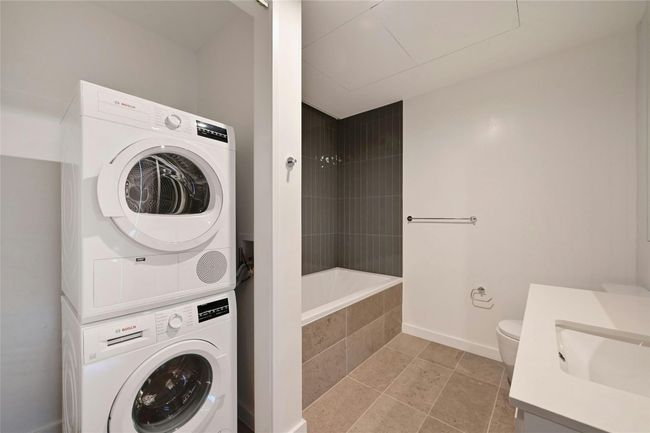 Sleek and functional bathroom featuring a modern soaking tub with vertical tile surround, neutral stone flooring, and a clean white vanity. A stacked Bosch washer and dryer set is conveniently located just off the bathroom, offering an efficient and space-saving laundry solution | Image 13