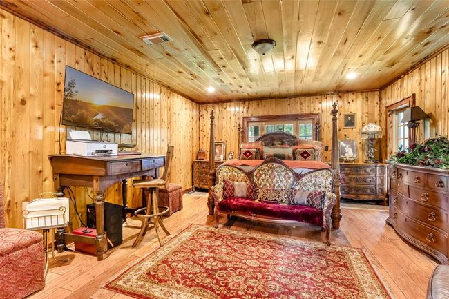 This cozy room features warm wood-paneled walls and ceiling, creating a rustic ambiance. It includes classic furniture with rich textures, a decorative rug, and a mounted TV. The space combines comfort with a touch of vintage charm, perfect for relaxation. | Image 33