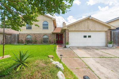 This is a two-story suburban home featuring a combination of brick and wood siding, with a prominent two-car garage and a well-maintained lawn. The home presents a welcoming red front door . | Image 2