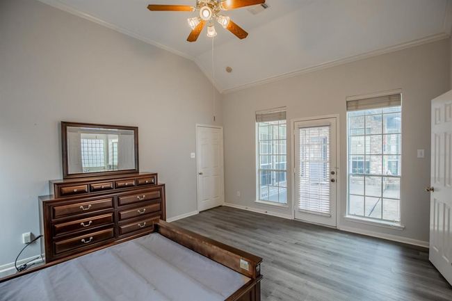 This spacious master bedroom features high ceilings, a ceiling fan, and large windows for ample natural light. It includes a door leading backyard, laminate flooring, and a classic wooden dresser with a mirror. | Image 28