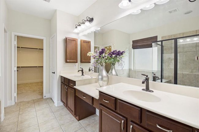 This is a spacious bathroom featuring a double vanity with cherry wood cabinets and elegant fixtures. It includes a large mirror, a glass-enclosed shower, and access to a sizable walk-in closet. Neutral tile flooring and soft lighting enhance the modern, clean look. | Image 16
