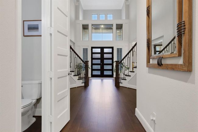 This photo showcases a modern, spacious entryway with high ceilings and abundant natural light. It features elegant dark wood flooring, a stylish front door with frosted glass panels, and a grand staircase with upgraded wrought iron railings. A sleek powder room is conveniently located to the side. | Image 26