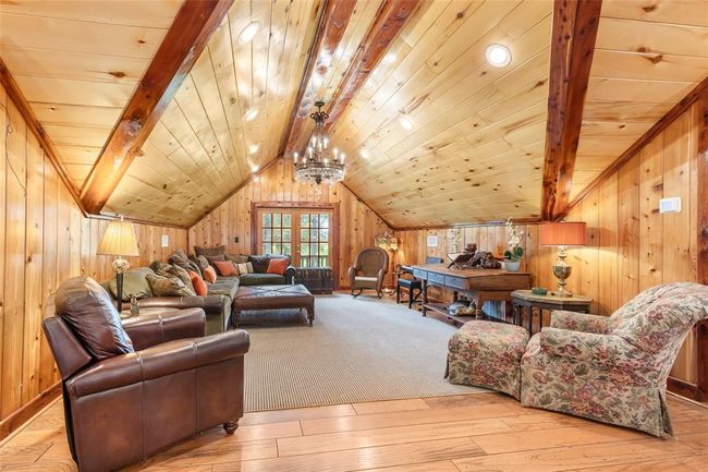 This cozy attic living space features warm wooden walls and ceiling with exposed beams, creating a rustic charm. It includes comfortable seating, a chandelier, and ample natural light from a windowed door, perfect for relaxation or entertaining. | Image 39