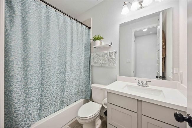 This bathroom features a clean, modern design with a light color palette. It includes a bathtub with a blue patterned shower curtain, a white vanity with a sleek sink, and a large mirror. A shelf above the toilet provides extra storage, accented with a small plant for a touch of greenery. | Image 21