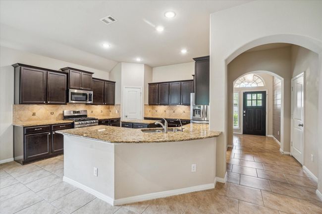 This kitchen features dark wood cabinetry, granite countertops, a central island, and stainless steel appliances. It offers a spacious layout with tiled flooring and recessed lighting, perfect for modern living. | Image 14